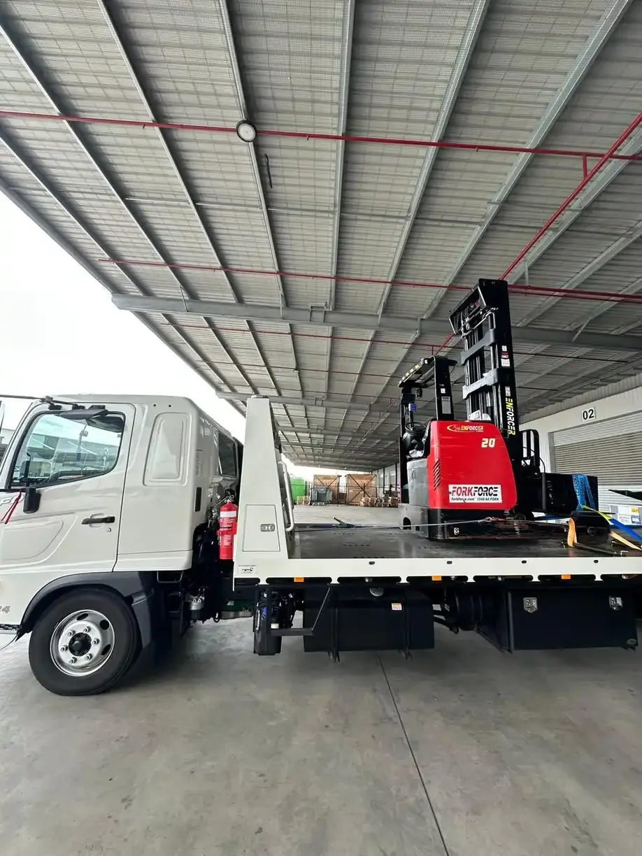 Red forklift strapped onto a tilt-tray truck under a warehouse canopy
